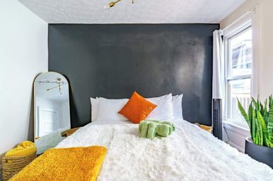 Cozy modern bedroom with a charcoal accent wall, fluffy white bed, orange accent pillow, yellow throw, green towels, leaning mirror and snake plant by a sunlit window.