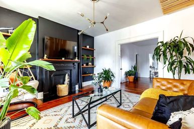 Bright modern boho living room with tan leather sofa, black fireplace wall and mounted TV, glass coffee table, patterned rug, hardwood floors, and large indoor plants