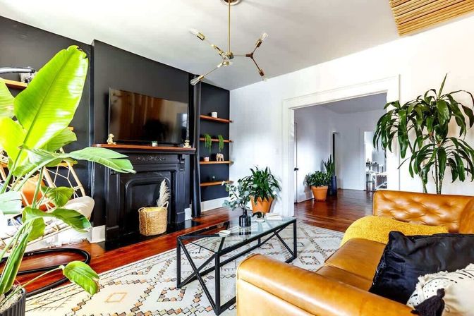 Bright modern boho living room with tan leather sofa, black fireplace wall and mounted TV, glass coffee table, patterned rug, hardwood floors, and large indoor plants