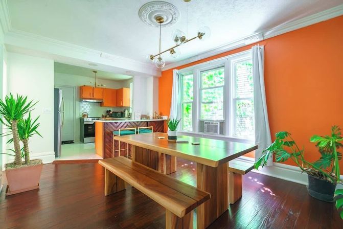 Sunlit open‑plan dining room with a bold orange accent wall, large windows, solid wood table and benches on dark hardwood floors, potted indoor plants, brass mid‑century light fixture, and a view into an orange‑cabinet kitchen.