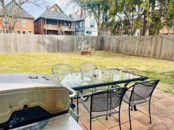 Cozy urban backyard with wooden privacy fence, grassy lawn, brick patio with glass-top table and metal chairs, gas grill and small fire pit, older houses visible beyond the fence.