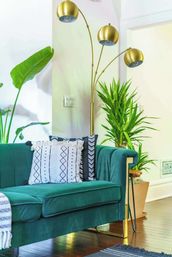 Emerald green velvet sofa with patterned throw pillows beside a gold triple-arched floor lamp and potted tropical plants on a hardwood floor in a bright modern living room