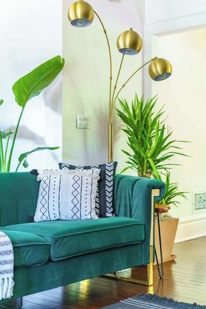 Emerald green velvet sofa with patterned throw pillows beside a gold triple-arched floor lamp and potted tropical plants on a hardwood floor in a bright modern living room