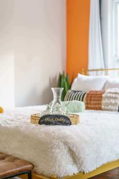 Sunlit cozy boho bedroom with white faux-fur bedspread, gold tray holding a glass carafe and glasses, black sleep mask reading "Plant Daddy", orange accent wall and houseplant