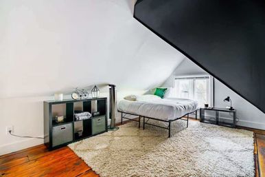 Cozy attic loft bedroom with sloped white ceiling, hardwood floors and large cream shag rug; minimalist metal-framed bed with white bedding and green accent pillow, black cube storage with bins and decor, and a small black nightstand and lamp by a window letting in natural light.