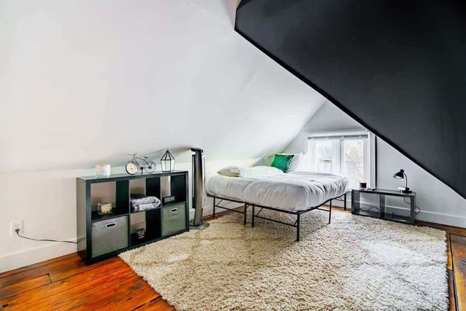 Cozy attic loft bedroom with sloped white ceiling, hardwood floors and large cream shag rug; minimalist metal-framed bed with white bedding and green accent pillow, black cube storage with bins and decor, and a small black nightstand and lamp by a window letting in natural light.
