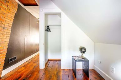 Bright loft-style attic nook with sloped white ceiling, exposed brick wall, wide-plank hardwood floors, open closet with hangers, and a small black side table topped with a globe and magazine.