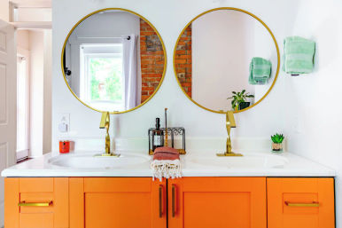 Bright loft-style bathroom double vanity with bold orange cabinets, white countertop, two round gold mirrors and brass faucets, green hand towels and small succulent plants.