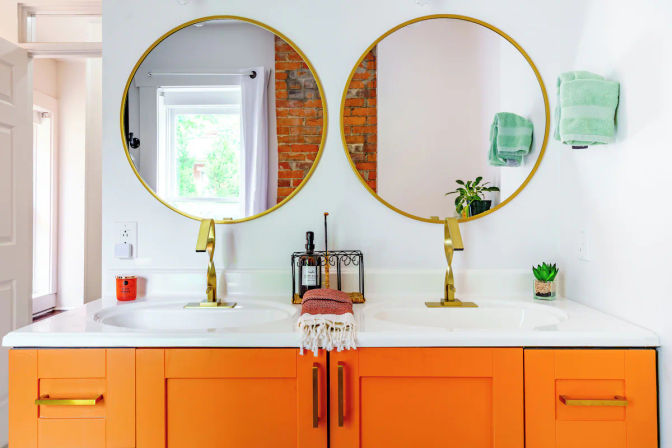Bright loft-style bathroom double vanity with bold orange cabinets, white countertop, two round gold mirrors and brass faucets, green hand towels and small succulent plants.