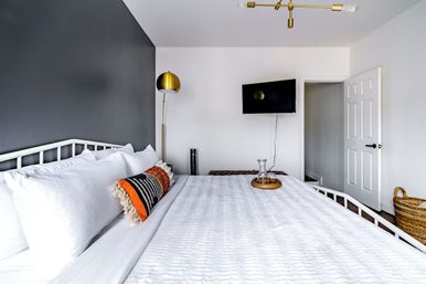 Bright modern bedroom interior with crisp white bedding, gray accent wall, brass floor lamp, patterned orange lumbar pillow, wall-mounted TV, open white door and wicker basket