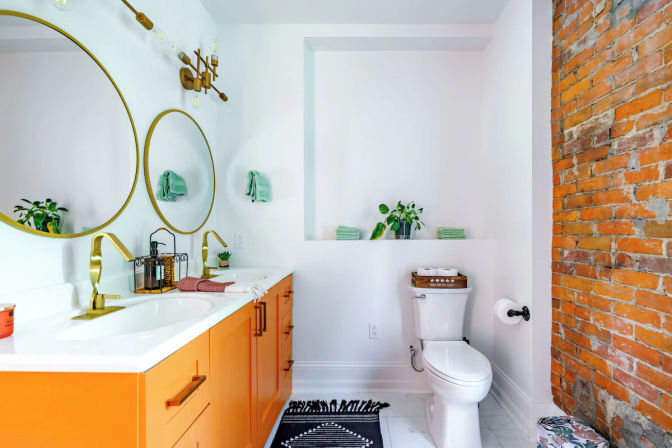 Bright modern bathroom interior featuring an orange double-sink vanity with brass faucets and two round gold mirrors, exposed red brick accent wall, white walls and marble floor, toilet, green towels, potted plants and a black patterned rug.