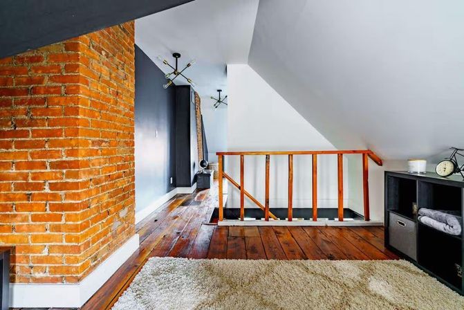 Cozy attic loft with exposed red brick wall, sloped white ceiling, polished hardwood floors, wooden stair railing, modern pendant lights and a shag area rug.