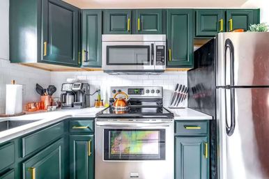 Modern apartment kitchen — dark green cabinets with brass pulls, white subway tile backsplash, stainless steel fridge, microwave and stove, copper kettle and coffee station.