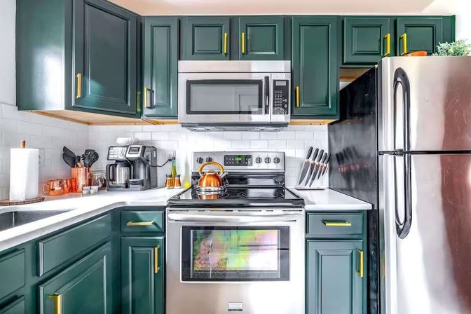 Modern apartment kitchen — dark green cabinets with brass pulls, white subway tile backsplash, stainless steel fridge, microwave and stove, copper kettle and coffee station.