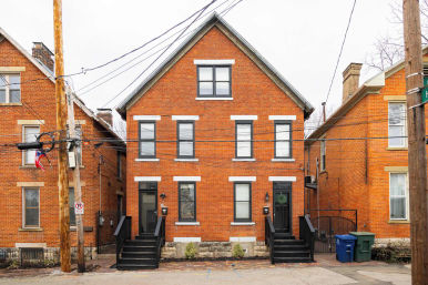 Symmetrical red brick duplex with black front steps and two black doors, white-trimmed windows, overhead utility wires and poles on an urban residential street with recycling bins.