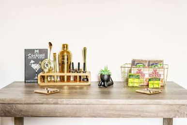 Bright minimalist wooden table styled as a mini home bar and snack station: gold cocktail shaker and bar tools in a wooden rack with a cocktail recipe book, a small potted succulent, and a wire basket holding instant noodle cups and green tea packets against a white wall.