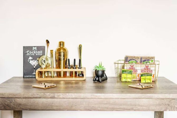 Bright minimalist wooden table styled as a mini home bar and snack station: gold cocktail shaker and bar tools in a wooden rack with a cocktail recipe book, a small potted succulent, and a wire basket holding instant noodle cups and green tea packets against a white wall.