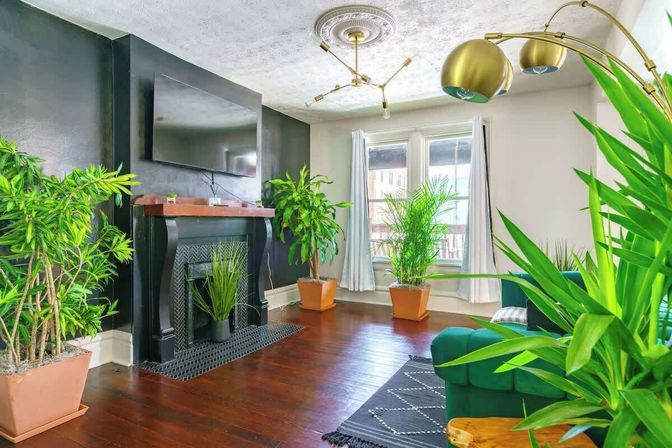 Sunlit modern living room with a black accent wall, decorative fireplace and wall-mounted TV, hardwood floors, emerald green sofa, brass light fixtures, and numerous potted houseplants by tall windows.