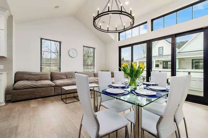 Sunlit modern open-concept living and dining room with glass-top table set for six, white chairs, yellow tulip centerpiece, black-framed sliding doors and transom windows, gray sectional sofa, round chandelier, and light hardwood floors.