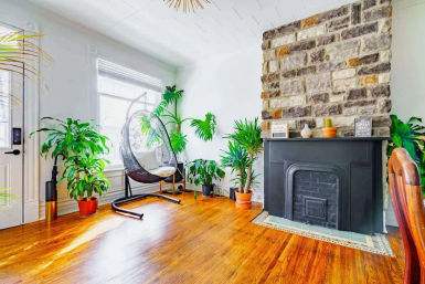 Sunlit, airy living room with a stone brick fireplace and black mantel, hanging egg chair, multiple potted indoor plants and warm hardwood floors — modern boho cozy vibe.