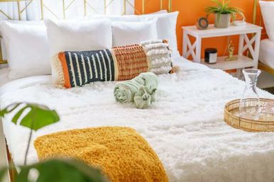 Cozy boho bedroom with fluffy white bedding, patterned lumbar pillows, mustard throw, rolled green towels and wicker tray with a glass carafe against an orange accent wall.
