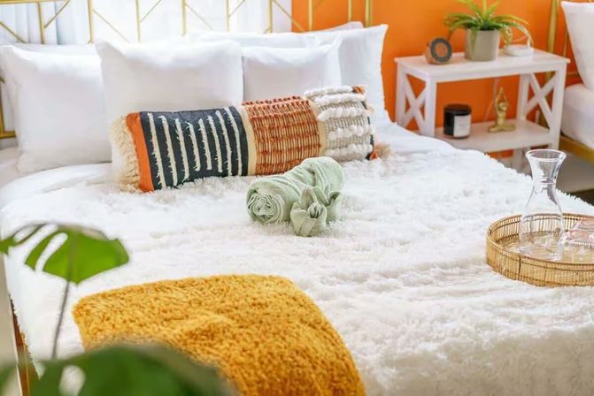 Cozy boho bedroom with fluffy white bedding, patterned lumbar pillows, mustard throw, rolled green towels and wicker tray with a glass carafe against an orange accent wall.