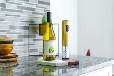 Gold electric wine opener and bottle of white wine on a marble-and-wood board atop a gray mosaic-tile kitchen countertop, stacked wooden bowls nearby.
