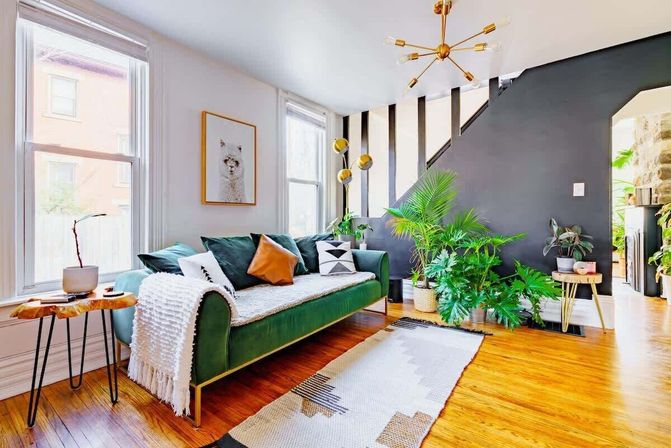 Bright modern living room with a green velvet sofa, gold mid-century chandelier, black accent stair wall, hardwood floors, patterned rug, large windows and lush potted plants