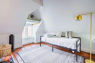 Bright cozy attic bedroom with sloped ceiling, white daybed, patterned gray rug, warm hardwood floors, gold floor lamp and sunny window seat.