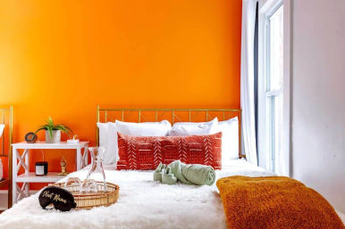 Cozy modern bedroom with bright orange accent wall, gold metal headboard, white fluffy bedding, rust-red patterned pillows and orange throw, white bedside table with potted plant and candle, tray with glassware by a sunlit window.