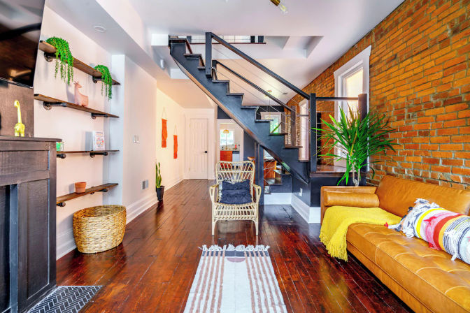 Cozy urban loft living room with exposed brick wall, dark hardwood floors, black metal staircase, rattan chair, tan leather sofa, colorful throws and potted plants.