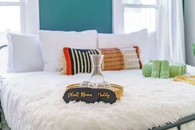 Cozy bright bedroom with white faux-fur bedspread, teal accent wall, woven and striped throw pillows, gold tray holding a glass carafe and glasses, green rolled towels, and playful “Plant Mama” and “Daddy” signs.