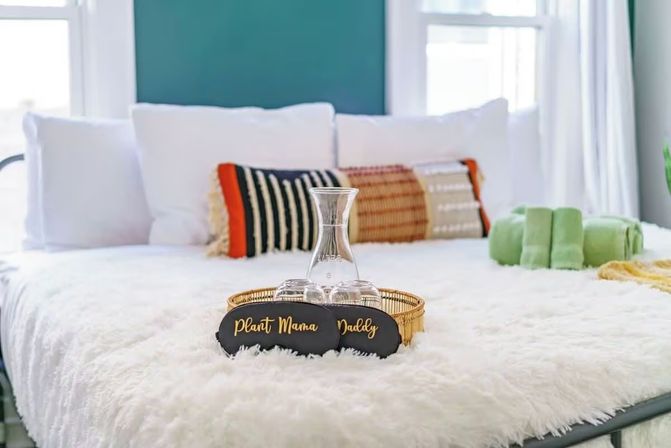 Cozy bright bedroom with white faux-fur bedspread, teal accent wall, woven and striped throw pillows, gold tray holding a glass carafe and glasses, green rolled towels, and playful “Plant Mama” and “Daddy” signs.
