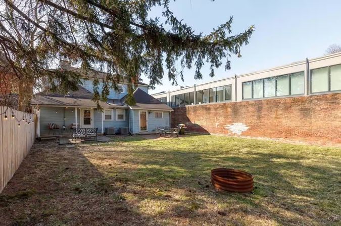 Sunny urban backyard with blue-sided house, grassy lawn, rusty metal fire ring, patio seating, string lights, and a tall brick wall with glass-fronted building beyond.