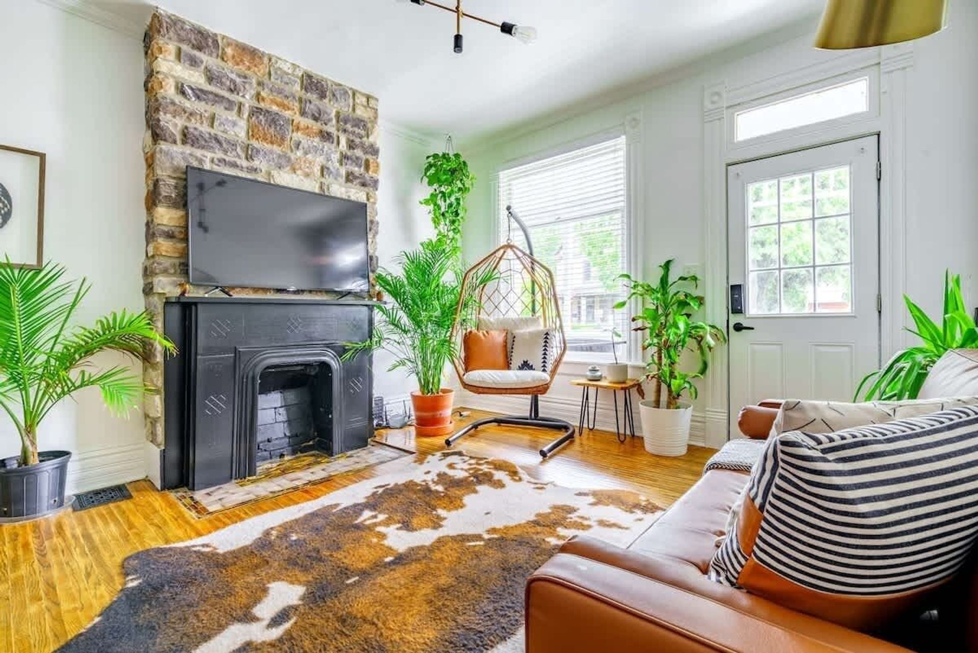 Sunlit cozy living room with a rustic stone fireplace and black mantel, wall-mounted TV, rattan hanging chair with cushion, abundant potted plants, cowhide rug and brown leather sofa on hardwood floors.