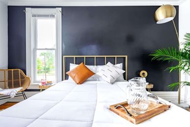 Cozy sunlit bedroom with charcoal accent wall, brass headboard, crisp white bedding, orange throw pillow, wooden tray with glass pitcher, rattan chair and brass floor lamp.