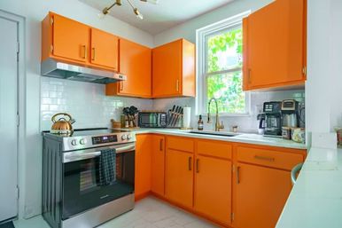 Cheerful L-shaped kitchen with bold orange cabinets, stainless steel range, white subway tile backsplash, gold faucet and a leafy window view.