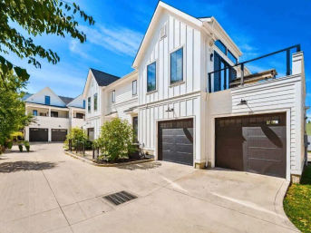 Modern white townhouses with vertical siding and black garage doors lining a shared concrete driveway, landscaped shrubs and glass-railed balconies under a bright blue sky