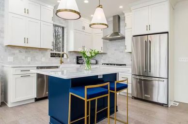 Bright modern kitchen with white shaker cabinets and marble countertop island in navy, two gold-framed blue bar stools, stainless steel French-door refrigerator, brass faucet, pendant lights, and light hardwood floors.