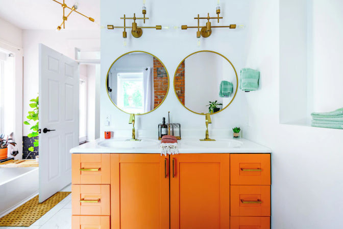 Vibrant modern bathroom with orange double-sink vanity, white countertop, two round gold mirrors, brass faucets and light fixtures, mint-green towels, potted plants, and exposed brick reflected in the mirrors.