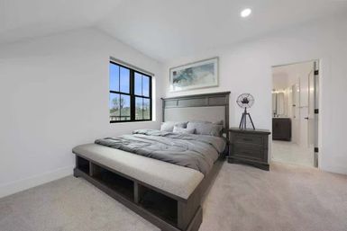 Cozy modern master bedroom with gray bedding, dark wood headboard and nightstand, upholstered storage bench, black‑framed window, and en-suite bathroom