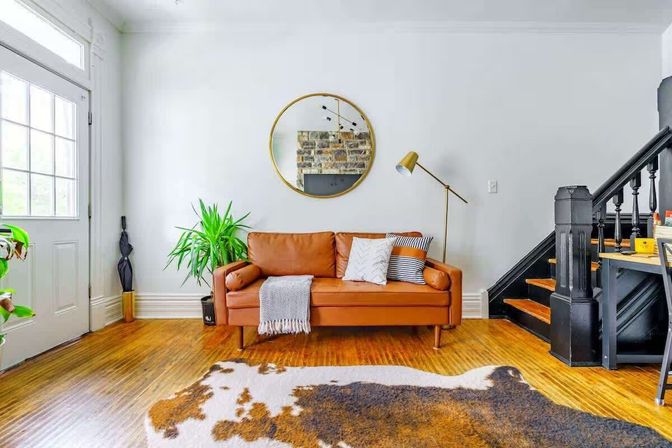 Bright modern living room featuring a tan leather mid-century sofa with throw pillows and a knit throw, round gold wall mirror, brass floor lamp, potted plant, cowhide rug on hardwood floors, black staircase, and a nearby front door.