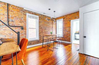 Cozy urban loft dining area with exposed brick walls, polished hardwood floors, tall white-trim windows, high wooden table and metal stools, glass-globe pendant light and orange accent chair.