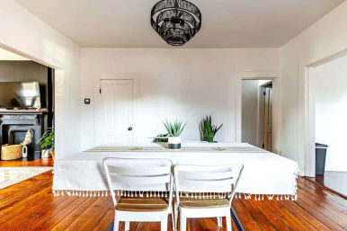 Bright minimalist dining room with white walls, long table draped in a fringed white tablecloth, two white chairs, warm hardwood floors, black wire chandelier, and potted plants near open doorways