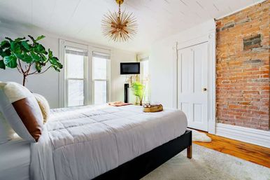 Sunlit cozy bedroom with white bedding, exposed red brick accent wall, hardwood floors, fiddle-leaf fig plant, and gold starburst chandelier.
