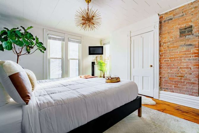 Sunlit cozy bedroom with white bedding, exposed red brick accent wall, hardwood floors, fiddle-leaf fig plant, and gold starburst chandelier.