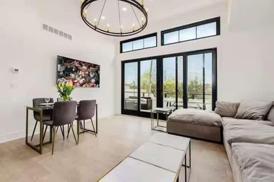 Bright modern living-dining room with a gray sectional, round chandelier, wall-mounted TV, and black-framed sliding glass doors opening to a sunny patio.