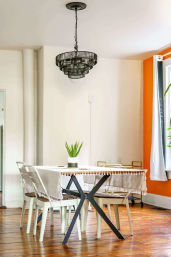 Bright modern dining room with wooden floors, X‑style black metal table base, white metal chairs and fringed tablecloth, small potted succulent centerpiece beneath a layered black wire chandelier by an orange accent wall and window.