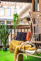 Cozy urban balcony oasis with rattan hanging chairs, mustard-yellow throws, black patterned and leather cushions, potted succulent, hanging pothos, and green turf flooring against a brick building backdrop.