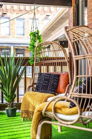 Cozy urban balcony oasis with rattan hanging chairs, mustard-yellow throws, black patterned and leather cushions, potted succulent, hanging pothos, and green turf flooring against a brick building backdrop.